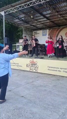 A wide outdoor shot of a stage set up with a musical band performing for an audience. There are five people on the stage. Three men are playing musical instruments, one on guitar, one on an accordion, and one on the drums which are partially visible. Two women are singing. The woman on the right is wearing a red dress, and the woman on the left is wearing a black dress. There's a white banner below the stage with a red "Крутий Заміс" logo and text in Ukrainian, and to its right, another banner with "Громадська органiзацiя Холодний Яр - Україна" written in Ukrainian. In front of the stage, a person with their back to the camera is raising their arms, seeming to participate in the music. The area in front of the stage is paved with concrete, and trees are visible in the background, suggesting an outdoor event.