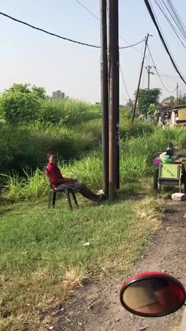 A young male wearing a red shirt and dark shorts is sitting on a black chair next to a utility pole. He is looking to his left and appears to be holding a phone in his right hand. The chair is positioned on a grassy area with tall green vegetation behind it. To the right of the utility pole, there is a small cart with a green container and what appears to be some food items. Further in the background, there are several makeshift dwellings and more vegetation. Overhead, power lines are visible. The foreground shows a gravel path and a red motorcycle mirror. The scene is illuminated by daylight, suggesting it is daytime.