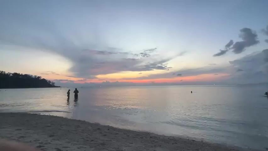 The scene displays a tranquil beach at dusk, with a few individuals enjoying the calm waters. The sky is adorned with soft, multicolored clouds, indicative of a sunset or sunrise. Two individuals are seen standing in the shallow water near the shoreline, while another person is floating further out. The beach consists of light-colored sand, and gentle waves are visible near the shore. A landmass covered with vegetation is present on the left side of the frame.