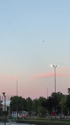 The video shows an airplane flying in a clear sky during dusk. The sky transitions from a light blue at the top to a soft pink and orange hue near the horizon. Below the sky, there are silhouettes of trees, streetlights, and some buildings. The airplane is visible as a small dark shape against the vast sky.