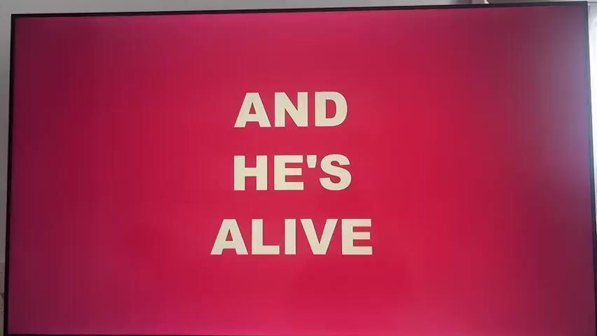 The video shows a television screen with a red background. White text appears in the center of the screen, displaying the phrases "AND HE'S ALIVE" and then "HALLELUJAH JESUS IS ALIVE." Music is playing in the background.