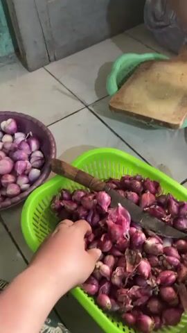 The video shows a close-up, slightly blurry view of a person's hands sorting through a large quantity of small red onions. The onions are in two containers: a purple bowl and a bright green plastic basket. A knife with a wooden handle rests in the green basket with the onions. In the background, there is a wooden cutting board on a green plastic mat and a tiled floor. The person's hands are actively reaching into the green basket, selecting and moving onions. Another red bowl is visible to the right. The overall scene appears to be a kitchen or food preparation area.