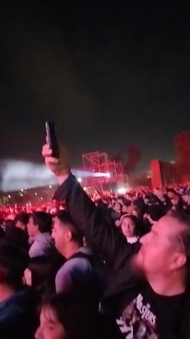 The video captures a large, densely packed crowd at a nighttime concert or event in Maipú, Chile. The subjects are numerous individuals, mostly seen from the side and back, extending far into the distance. Many attendees are holding up smartphones, seemingly recording or photographing the event. The foreground shows people in casual, dark attire, with one person wearing a black jacket with a white pattern on the shoulder.

The scene is dominated by stage lighting, which emits strong blue, white, and red beams of light that pierce through clouds of smoke or fog, creating an energetic atmosphere. The stage itself is not directly visible, but its presence is indicated by the light and smoke effects. The activity centers around the crowd watching and documenting a performance. The time of day is clearly night. No visible text is legible within the video frame.