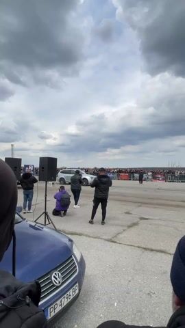 The video captures a drag race between two vehicles on a concrete road. On the left side of the frame, a blue Volkswagen car is partially visible. The main focus is on the two vehicles lined up for the race: a Mitsubishi Pajero SUV and a silver Audi station wagon. A crowd of spectators is visible behind a fence, watching the race, and several people are standing near the starting line, including photographers. The Pajero initially loses traction, kicking up dust as it accelerates down the road. The sky is overcast with dark clouds. A large speaker is positioned on a stand to the left of the road.