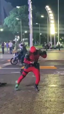 A person dressed in a Deadpool costume is dancing in the street at night. Several other people are standing in the background. There is a motorcycle parked nearby. A light post is visible in the background.