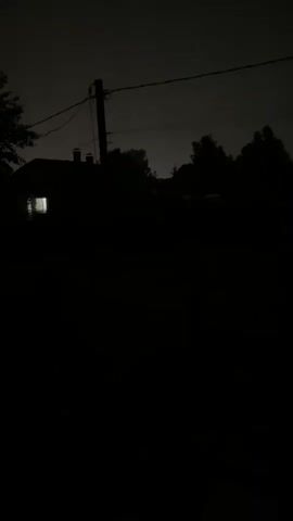 A nighttime view of a dark, rural setting. A single window of a building, likely a house, is illuminated from within, casting a faint glow. In the background, silhouetted trees and a utility pole with wires are visible against a dark sky. A fence runs horizontally across the middle ground, further obscured by darkness. The foreground is entirely black, suggesting deep shadow or an obscured area. No people or animals are visible.