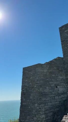 The camera pans from a portion of a light gray brick wall to a bright blue sky and a tranquil, light turquoise ocean. The sun shines brightly in the sky. The ocean appears flat and calm, with a slight glimmer on the surface. A single bird flies across the horizon. The edges of a cliff with some vegetation can be seen on the left and right edges of the frame.