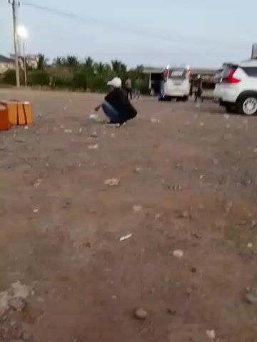 A person in a white hat and dark clothing squats in a dusty outdoor area, facing away from the camera. To their left, another person in an orange shirt and jeans stands looking down. In the background, several vehicles, including a white van and a white SUV, are parked near a low building. Palm trees and utility poles are visible in the distance. The ground is covered in gravel and debris.