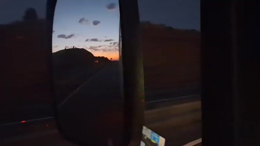 The video shows a view through a vehicle's side mirror, capturing a twilight scene. A road stretches into the distance, bordered by hills. The sky displays hues of orange and blue, with scattered clouds. A traffic signpost is visible in the distance. The lighting suggests it is either dawn or dusk.