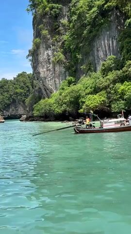 A longtail boat is partially visible in the turquoise water, with a person in a yellow shirt and a red vest standing on it. The boat has a long wooden outrigger extending from its side. Behind the boat, a steep, rocky cliff face covered in lush green trees rises dramatically from the water. The sky above is blue with some clouds. The water is clear and reflects the surrounding greenery.