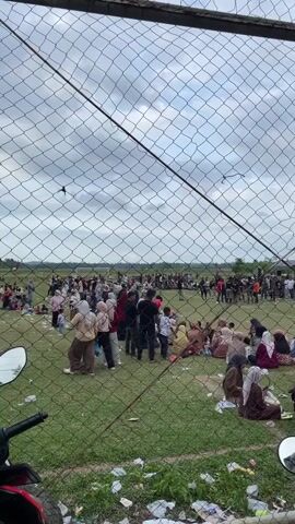 The video is filmed from behind a chain-link fence, looking out onto a grassy field filled with people. The sky is cloudy. Many people are sitting or standing on the grass, some in small groups, others in larger crowds. They appear to be watching something happening in the distance, where there is a line of people standing in what looks like a race or competition. Some people are wearing hijabs. There is some litter on the ground in the foreground, and a motorcycle is partially visible on the left side.
