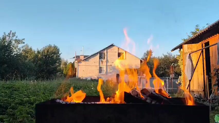 The video shows a close-up of a fire burning in a metal barbecue grill. Flames lick upwards from burning logs. In the background, a two-story house with brick siding is visible, bathed in the warm light of the sun. To the right of the house is a wooden shed. Surrounding the house and shed is lush green foliage, suggesting an outdoor setting, possibly a backyard or garden. The sky above is a clear blue. The time of day appears to be late afternoon or early evening, based on the quality of the light. There are no people or animals visible in the frame, and no explicit actions are taking place beyond the burning fire. The overall impression is one of a peaceful, rural or suburban scene at dusk.