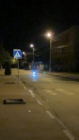 A person rides an electric scooter at night on a street. The scooter has blue and white lights. There is a pedestrian crossing on the street and a pedestrian crossing sign. The person on the scooter is looking at the camera and smiling. The street is lit by streetlights and the lights from the scooter.