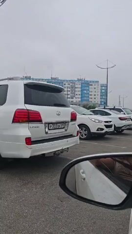 The video shows a parking lot on a cloudy day. Several white cars are parked in a row, including a Lexus SUV in the foreground. In the background, a multi-story building with blue and white accents is visible. The camera appears to be inside another vehicle, as a side-view mirror is in the bottom right corner of the frame, reflecting part of the interior and the car it belongs to.