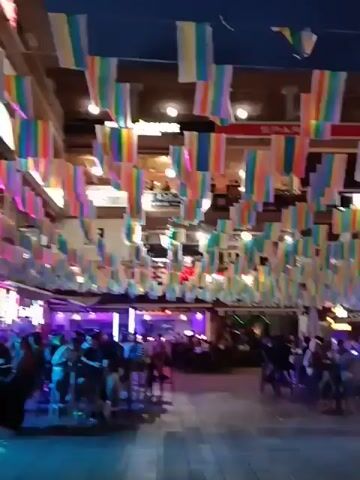 The camera pans across a vibrant outdoor nightlife scene. Overhead, rows of multicolored striped flags adorn the ceiling, adding to the festive atmosphere.  A large crowd of people, mostly seated, populate the area, likely patrons of the surrounding bars and restaurants. In the background, the "Blue Havana" establishment is clearly visible. Overall, the scene captures a lively and bustling outdoor gathering.