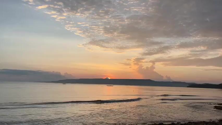 The video shows a sunrise over the ocean. The sun is a small orange disc just above the horizon, casting a warm glow across the sky and water. The sky is filled with scattered clouds, some tinged with pink and orange from the rising sun. The ocean is calm, with gentle waves lapping at the shore. In the distance, a dark silhouette of land can be seen, with a few small boats dotted on the water. The foreground shows a rocky shore covered in seaweed. The overall atmosphere is peaceful and serene.