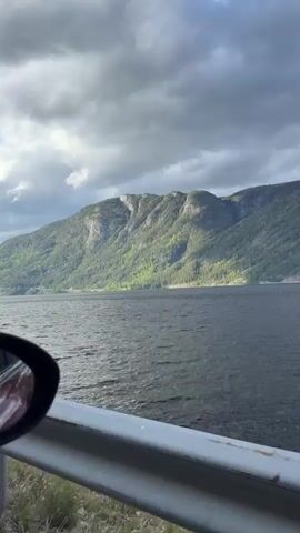 The video shows a car traveling along a road next to a lake surrounded by mountains. The sky is cloudy with patches of blue sky. The video is taken from the passenger seat and shows the side mirror and the road barrier.
