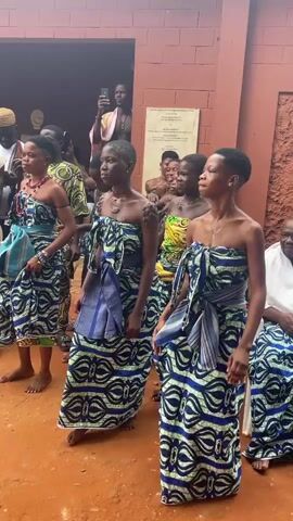 This is a medium shot video of a group of people, mostly young women, dancing. The women are wearing matching blue and white patterned strapless dresses with sashes tied around their waists and shoulders. They are barefoot and appear to be in an outdoor courtyard with a red-brown dirt floor. Behind them is a building with a reddish-brown brick facade and a dark opening, possibly a doorway or entrance. Several people, predominantly men, are standing or sitting in the background, some of whom appear to be filming the dancers with their phones. The lighting suggests it is daytime. The overall atmosphere appears to be celebratory or ceremonial.