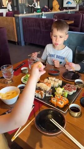 A group of people are seated at a wooden table in a restaurant, eating sushi. A young boy in a grey t-shirt with the words "SHINE BRIGHT" is in the center of the frame. He is holding chopsticks and appears to be eating. To his right, a young person in a black and white striped sweater is also holding chopsticks and drinking from a glass. To the boy's left, another person, mostly out of frame, is holding chopsticks and reaching for food. In front of the boy is a large platter of sushi, various small bowls of sauce and condiments, and a bowl of french fries. The restaurant appears to be upscale, with dark purple upholstered seating and dim lighting. In the background, other diners and staff can be seen. The camera is positioned at table level, looking slightly down at the food and people. The overall atmosphere is casual and familial.