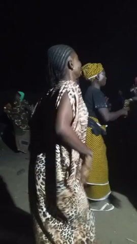 The video shows a group of women dancing outdoors at night. The woman in the foreground is wearing a long, loose-fitting dress with a leopard print pattern. The other women are wearing a variety of colorful clothes, including yellow and green patterns. They are moving to a beat, raising their hands. It is dark, and the lighting is dim. The ground appears to be sandy or dirt.