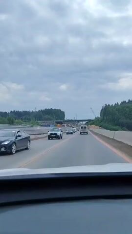 The video shows a car's perspective driving on a highway with multiple lanes. Other cars are present, including a white van directly ahead. The road has orange lines marking the edges. In the distance, there's a bridge and construction equipment, including a crane. The sky is overcast. The camera appears to be dashboard-mounted in the driver's seat.