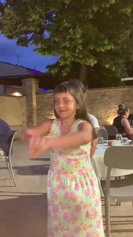 A young girl in a floral dress dances joyfully outdoors at dusk. She is in the foreground, smiling and moving her arms energetically. Behind her, another girl in a white t-shirt and a silver skirt is also dancing. They appear to be at an outdoor restaurant or event with tables set with white tablecloths and chairs visible. Trees and a brick wall are in the background, and the sky is a deep blue.