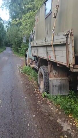 A medium shot, taken from the side of a rural road, shows the rear of a weathered, olive-drab truck with a wooden flatbed. The truck is parked on the side of the road, partially on the grass. The rear of the truck's cargo area, which appears to be a modular unit with a white cross on a red circle visible, is in the foreground. A license plate with Cyrillic characters and the number Y891TH799 is visible. The background consists of trees and foliage, suggesting a wooded area. The road surface is asphalt and appears to be in disrepair. There are no people visible in the frame.
