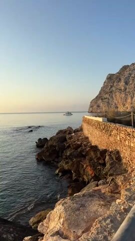 A drone shot pans slowly across a rocky coastline. The sun is low in the sky, casting a warm glow on the scene. The sea is calm, with a few rocks visible near the shore. A white boat is visible in the distance on the water. A stone wall runs along the top of the cliff, with a rope fence. The sky is clear and blue.