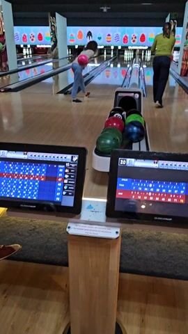 Here's a description of the video:
The video shows two people bowling at a bowling alley. The camera is positioned behind the ball return. The person on the left throws a pink bowling ball down the lane, hitting several pins. A woman in green is preparing to bowl. The bowling alley is decorated with Easter-themed graphics. There are two scoring monitors in the foreground displaying scores. Several bowling balls of different colors sit in the ball return.