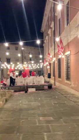 A night scene in an outdoor plaza, likely a European town square, with a building on the right side of the frame. The building has a light pink facade with several windows that are barred. Across the plaza, illuminated by strings of incandescent bulbs, a long white-draped table is set for a meal. On a raised platform in front of the table, several individuals in red shirts and white pants are standing. Some people are seated at the table. Red and white flags are strung along the building on the right. The ground is paved with cobblestones, and rose petals are scattered across the plaza, especially on the cobblestones in front of the raised platform.