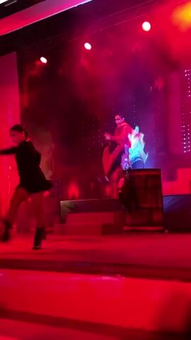 Two performers are on a red stage under red and purple lights with smoke effects. The performer on the left is in a dark outfit with buttons down the front and dark boots, and is crouched down with arms extended. The performer on the right is wearing a yellow shirt and dark pants, and appears to be seated on a barrel. They have a cape or sheet draped around them. Two wooden barrels are visible on the stage, and stairs are behind the performer on the right. The background appears to be a stage set with a dark curtain and lights. The video is in landscape orientation.