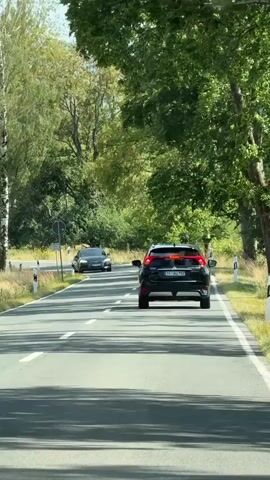 A black Mitsubishi Eclipse Cross drives on a paved road, followed by a black Audi. The cars are driving on a two-lane road with white lines. Trees and foliage are on both sides of the road. The sky is bright and the sun is shining.