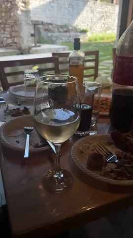 The video shows a close-up of a wooden table set for a meal. There are several plates with food remnants, glasses filled with drinks, a bottle of wine, and a large bottle of soda. A fork rests on one of the plates. In the background, through a large glass window, there are buildings and people. One man in a black t-shirt is standing and looking towards the table. The overall atmosphere suggests a casual outdoor gathering or meal.