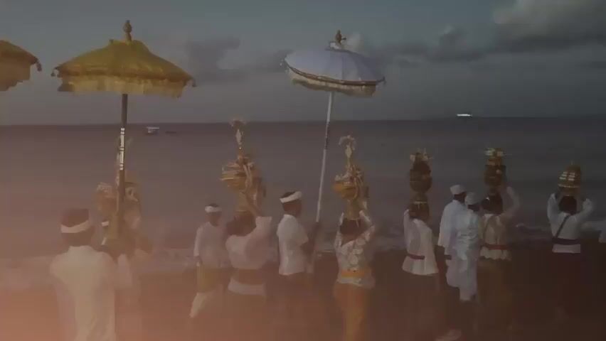 A group of people, dressed in traditional white clothing and head coverings, walk along a beach carrying offerings. They are walking towards the ocean, which is visible in the background. Some individuals hold ceremonial umbrellas above their heads. The sky is dark, suggesting it is either dawn or dusk. The scene appears to be part of a religious or cultural procession.