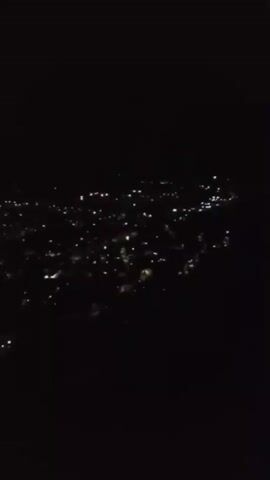 The video captures a nighttime aerial view of a city illuminated by numerous lights. To the right of the frame, a large, white cross stands out against the darkness. The camera perspective suggests it might be from an airplane or a drone. The lights of the city are spread out below, indicating a populated area. The cross appears to be on higher ground or a prominent location.
