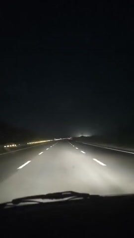 The video captures a first-person perspective of driving on a highway at night. The primary subject is the road ahead, illuminated by the vehicle's headlights, with white lane markings clearly visible. The highway appears to be a multi-lane road, with a median strip separating the lanes. On the left side of the road, a line of yellow reflectors or lights runs along the shoulder, suggesting a rural or less developed area. In the distance, faint lights of other vehicles and possibly streetlights can be seen, indicating ongoing traffic and infrastructure. The sky is dark, characteristic of nighttime, and there is no visible weather interference. The overall scene conveys a sense of solitary travel through a dark landscape, with the focus on the path forward. There are no people or specific objects other than the road and the immediate car interior (windshield and wipers). The time of day is clearly night. The location cues, such as the yellow reflectors and the general appearance of the highway, align with a setting in Araul, India. The emotion conveyed is one of steady movement and perhaps a touch of quiet introspection due to the solitary nature of the drive and the darkness. No text is visible in the media file.