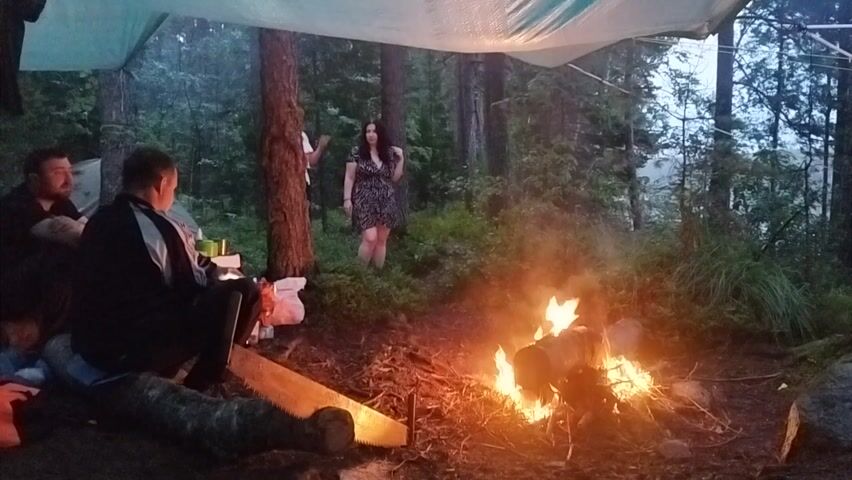 In a forest clearing, four individuals gather around a crackling campfire beneath a tarp shelter. The scene appears to be set in the evening or dusk, with the dim light filtering through the trees. Two men are seated in the foreground, one facing away from the camera, observing the fire, while the other is turned towards the camera, looking relaxed. Two women are standing further back, one appearing to converse with the other who is looking towards the fire. The setting is a natural, wooded environment, with trees, undergrowth, and a rough, earthy ground. The presence of a tarp suggests a campsite or an outdoor gathering where protection from potential rain or elements is provided. The fire is the focal point, casting a warm glow on the surroundings and the faces of those near it. The overall atmosphere seems casual and convivial, indicative of a social gathering in nature. No text is visible in the image.