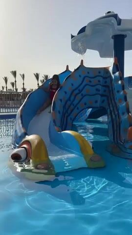 A young girl is playing on a water slide in a pool. She is wearing a red swimsuit with the words "bae-water" on it. The water slide is blue and yellow and shaped like a sea creature. The pool is a bright blue with palm trees visible in the background. The girl slides down the slide, splashing into the water, then stands up and smiles at the camera.