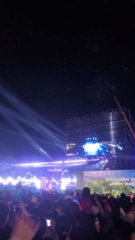 The video shows a large crowd of people at a nighttime event, likely a concert or festival. A stage is visible, adorned with bright lights and a large screen displaying imagery. Fireworks explode in the sky above, illuminating the scene. People in the crowd are holding up their phones, presumably recording the event. The sky is dark, but the lights from the stage and fireworks provide ample illumination.