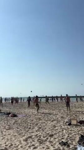 A wide shot shows a large group of shirtless people playing a game on a beach. They are spread out across the sandy beach in front of the ocean on a sunny day under a bright blue sky. The game involves throwing and kicking a ball. Some beach towels and bags are scattered on the sand in the foreground.