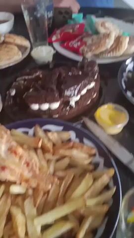 The video shows a table laden with food, likely a meal or a celebration. There are plates of meat skewers, french fries, a chocolate cake, pastries, and possibly some kind of dip or sauce. A glass of liquid is also visible. The camera pans across the table, focusing on different dishes. The lighting is somewhat dim, suggesting an indoor setting. The overall impression is one of abundance and a shared meal.