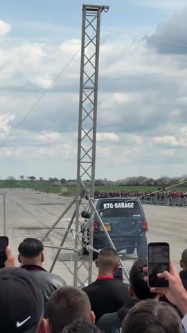 The video shows a dark-colored SUV with "RTG GARAGE" written on its back as it accelerates on a dusty track. Smoke comes out of the exhaust pipe. The car drives down the track, passing a person walking on the side and another vehicle waiting. The SUV is viewed from behind a crowd of spectators holding phones, and a metal tower structure is in the foreground. The sky is cloudy and there is a green field in the distance with some trees.