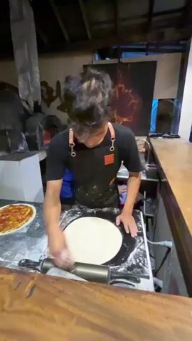 The video shows a busy kitchen environment where pizzas are being prepared. In the foreground, a person wearing a black apron is seen pressing dough with their hands. Next to them, a rolling pin lies on the counter. Further back, two individuals in blue shirts are actively involved in making pizzas. One person is spreading sauce on a pizza base, and another is adding toppings. In the background, a large stone oven with a fire burning inside is visible, indicating a traditional pizza-making setup. The overall scene suggests a lively and functional pizza-making operation.