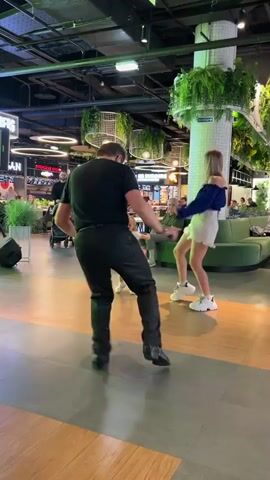 A man and a young woman are dancing in what appears to be a modern indoor space with a lot of greenery. The man, facing away from the camera, wears a black t-shirt and black pants with cowboy boots. The woman, also facing away, is wearing a blue off-the-shoulder top and a white skirt with sneakers. They are on a polished wooden floor, with large green lounge seating and cylindrical pillars adorned with hanging plants visible in the background. The setting suggests a public area, possibly a mall or a food court, given the presence of other people in the distance and what looks like signage for food establishments. One sign in the background appears to read "SOSTI ITALIANO." The lighting is bright and artificial, indicating an indoor environment during the day or evening. There is a sign with Russian text that appears to read "ВЫХОД," meaning "EXIT." The overall atmosphere seems lively and energetic, with onlookers observing the dance.