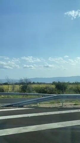 The video shows a view from a moving vehicle, looking out at a rural landscape. The sky is blue with scattered white clouds. In the middle ground, there is a field of tall green crops, possibly corn, bordered by a metal guardrail. Several trees are visible along the roadside, some bare and others with foliage that appears to be rustling in the wind. In the background, a range of hills or mountains is faintly visible against the horizon. The foreground shows a portion of a paved road with white lane markings. The overall impression is one of travel through a countryside on a sunny day.