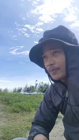 A man in a dark bucket hat and a dark zip-up jacket looks to his right while positioned low in the frame. He has dark hair and a light mustache. Behind him are patches of green grass and dry ground, with a line of trees or bushes in the distance under a bright blue sky with scattered white clouds. The man remains still, observing something off-camera.