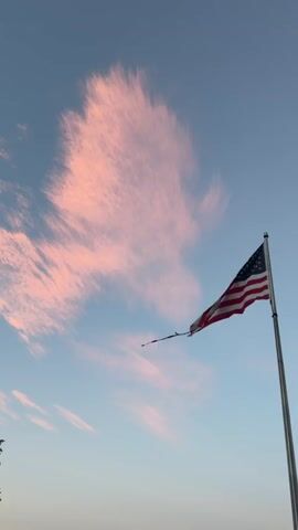 The video shows an American flag attached to a flagpole. The flag is red, white, and blue with stars and stripes. The flag is flapping in the wind. The sky is a light blue color and there are some pink clouds. The video is shot from a low angle, looking up at the flag and the sky.