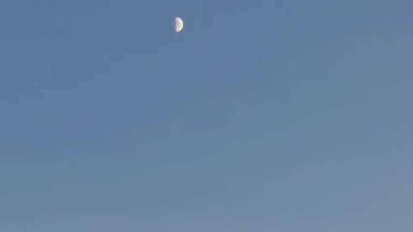 The video begins with a clear blue sky and a half-moon visible. The scene then transitions to a bright, hazy, light yellow sky. The last portion of the video shows a sunset with the sun low on the horizon, casting a warm orange glow. Trees and bushes are silhouetted against the sky on the right side of the frame. A road with white lane markings is visible curving to the left. A truck is driving on the road in the distance. A speed limit sign indicating 130 is visible on the right side of the road. The camera appears to be inside a moving vehicle.