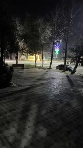 The video shows a night view of a park with a paved walkway.  The walkway is patterned with grey and white stones. The lighting is dim, and shadows from the bare trees are cast on the walkway.  Across the street, illuminated decorative lights are visible, including a glowing heart shape and a colorful, multi-pointed star shape.  A bench sits on the grass near the walkway. Cars pass by on the road in the background. A dark-colored car is parked on the side of the road.  Small patches of snow are visible near the walkway.  The scene has a quiet, slightly desolate atmosphere.