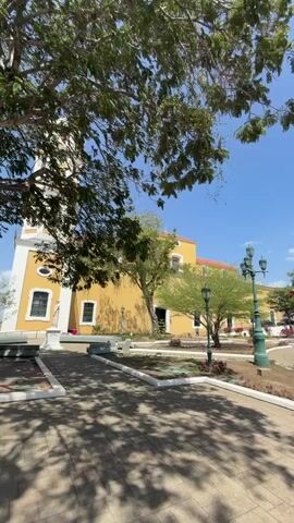 The video depicts a wide-angle view of a town square. The square has a statue in the center. It is surrounded by buildings of varying colors, including yellow, white, and pink. The sky is blue with scattered clouds. Several trees are present, some casting shadows on the paved ground. Lampposts line the perimeter of the square. There are a few people visible in the background, walking around. The overall environment suggests a sunny day in a historical or cultural area.